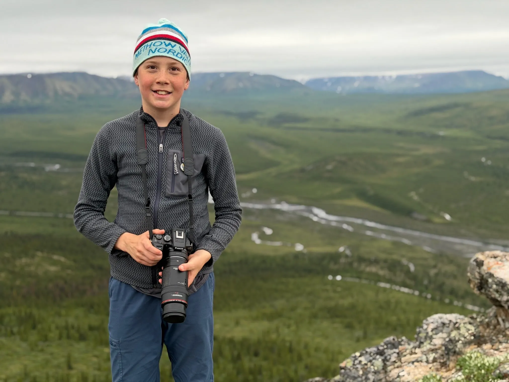 Jude Polson standing on a ridge above a wide river valley, holding a camera with a telephoto lens